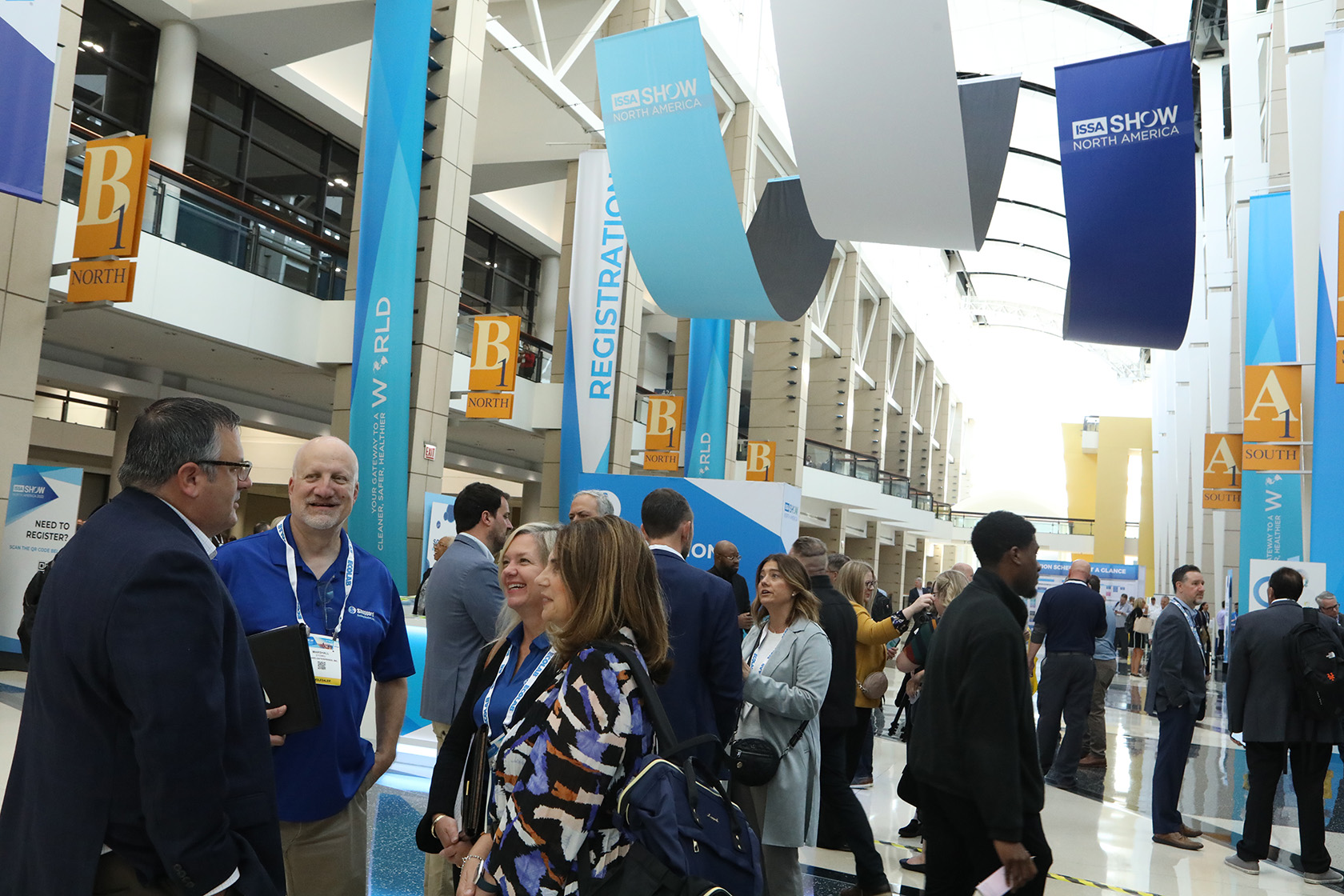 There is a group of people talking in the foreground while others are walking around in the background. ISSA Show North America banners can be seen above them hanging from the ceilings.
